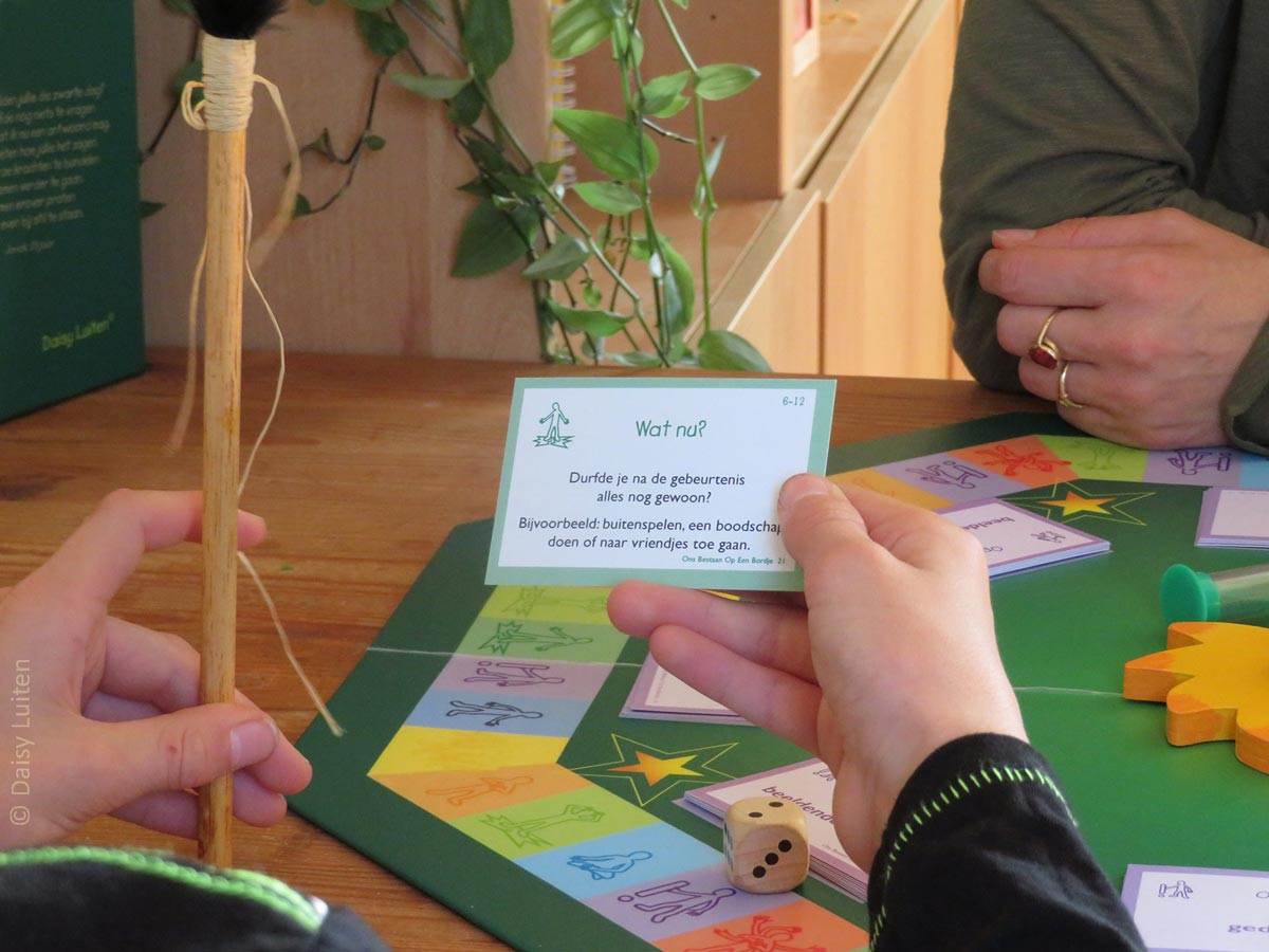 Wij kijken mee over de schouder van een kind. Hij heeft een vragenkaart in zijn hand uit de categorie 'Wat nu'. Op de achtergrond is het groene spelbord van Ons Bestaan Op Een Bordje zichtbaar met daarop een traject van kleurige vlakjes, een houten zon in het midden en een dobbelsteen. Op het spelbord liggen een aantal stapels paarse kaartjes.