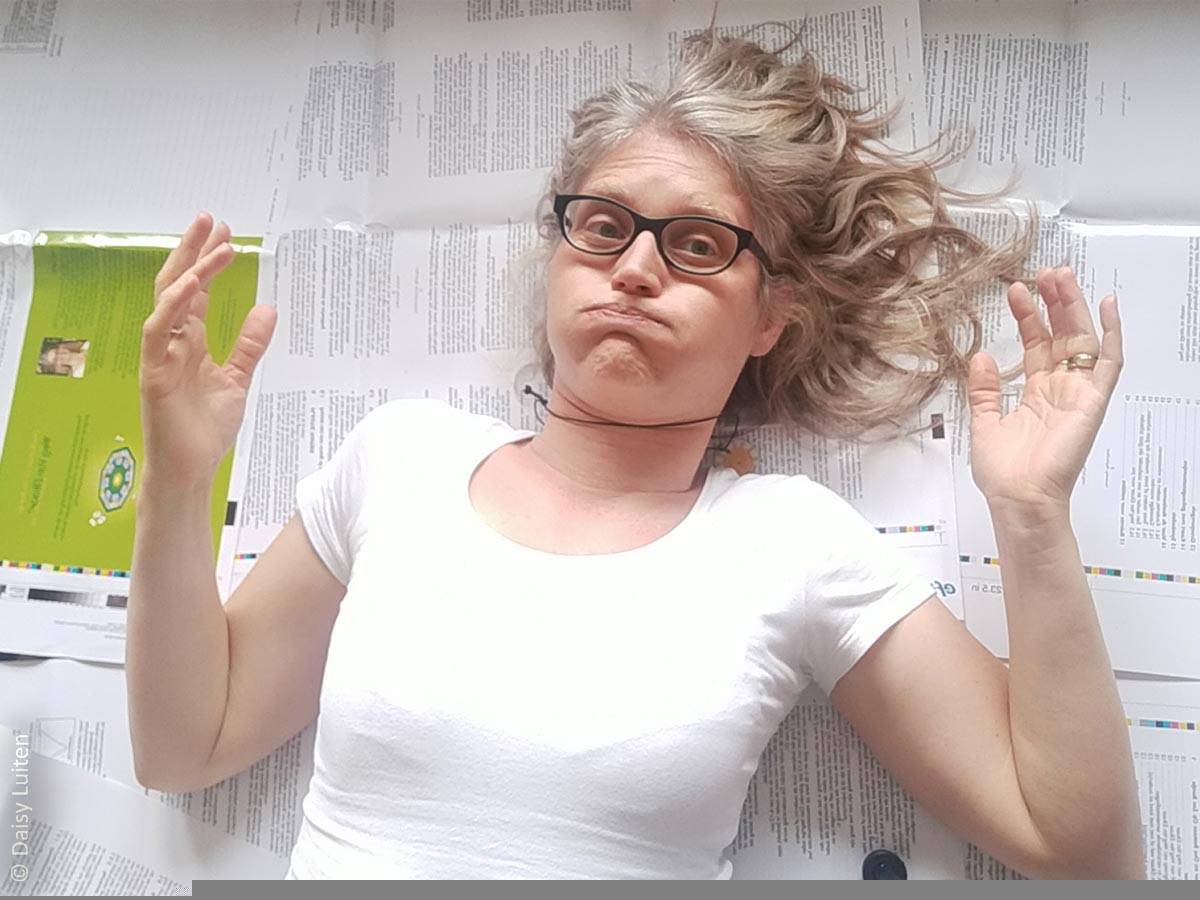 Daisy Luiten tijdens de ontwikkeling van Ons Bestaan Op Een Bordje. Ze ligt op drukvellen van haar tool waarop veel tekst te zien die gecontroleerd moet worden. Ook is de omslag van een van haar boekjes bij het therapeutische spel zichtbaar. Daisy heft haar handen omhoog en kijkt met een uitdrukking dat ze het aardig zat is en er zo een dikke zucht gaat volgen.