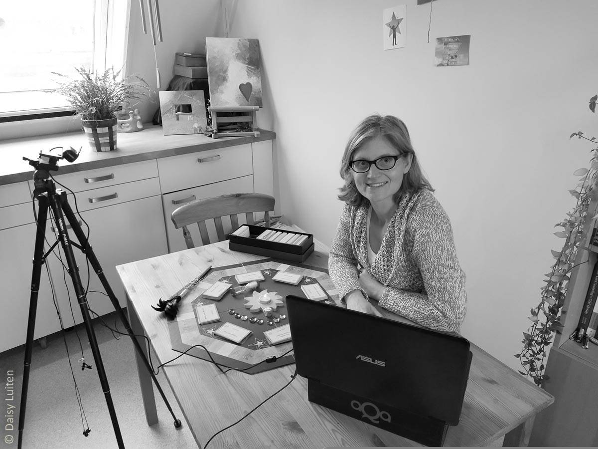 In een zwartwit afbeelding zien we Daisy Luiten aan tafel zitten met haar therapeutische tool uitgestald. Een camera op een statief staat op het spel gericht. Daisy heeft ook haar laptop voor zich waarop ze via beeldbellen online sessies geeft aan cliënten met Ons Bestaan Op Een Bordje, of aan therapeuten om hen te leren hoe met de tool te werken. De therapeutische tool die vertellen over een nare of pijnlijke ervaring vergemakkelijkt.