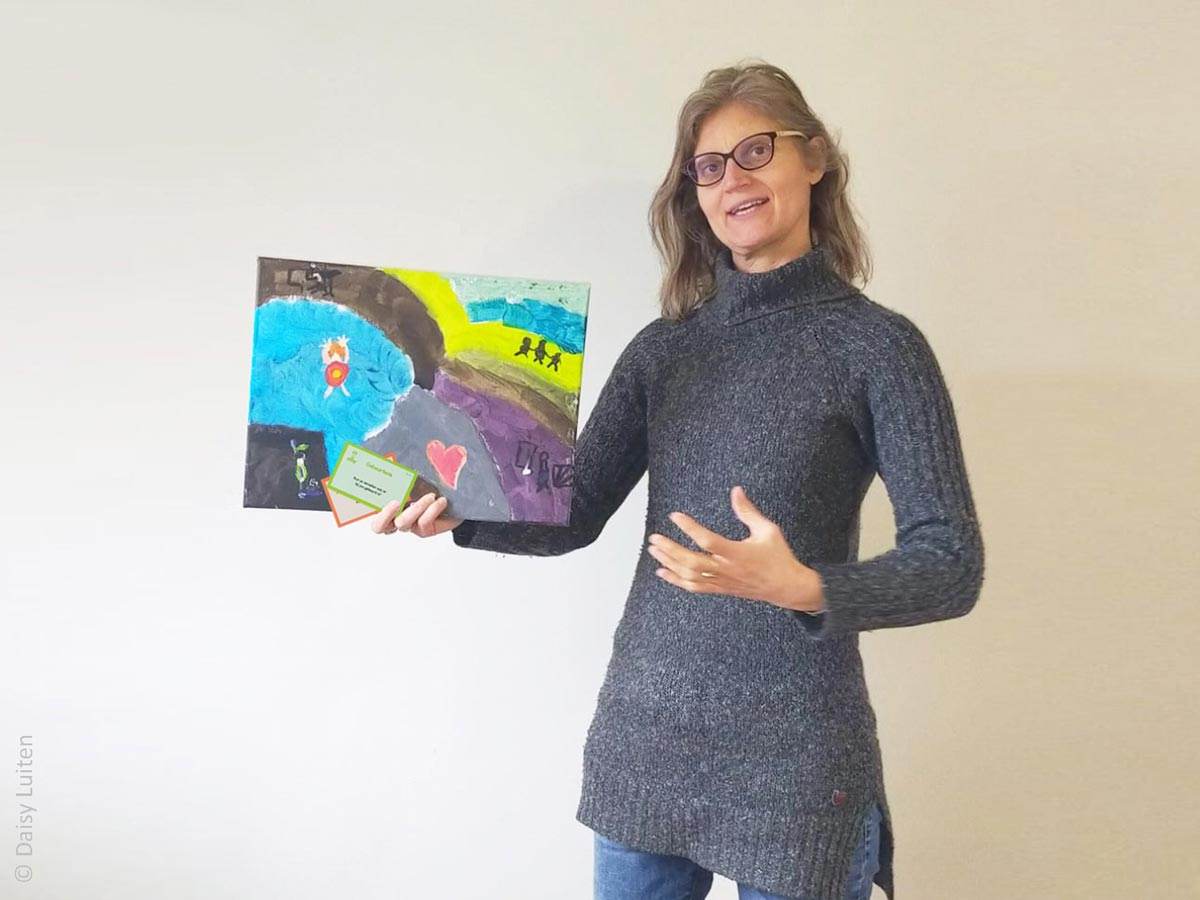 Daisy Luiten, een vrouw met donkerblond halflang haar en een bril, houdt tijdens het vertellen een schilderij en wat vragenkaartjes in de hand. Daisy geeft hier de creatieve verdiepende training rond haar tool. Een inspirerende training waarbij je in één dag fijne nieuwe werkvormen ontdekt. Om verrassend meer uit Ons Bestaan Op Een Bordje te halen.